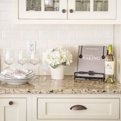 a kitchen counter with wine glasses, plates and flowers on top of it in front of white cabinets