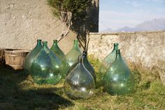 four green glass vases sitting in the grass near a wall and basket on the ground