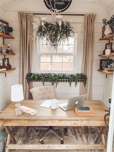 a desk with a laptop on it in front of a window filled with potted plants