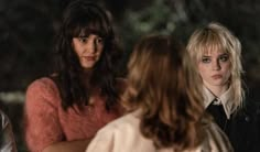 three women standing around talking to each other in front of some trees and one is wearing a pink sweater