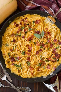 a skillet filled with pasta and bacon on top of a wooden table next to squash