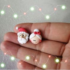 two tiny santa and mrs claus earrings on someone's hand with christmas lights in the background