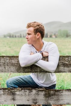 a young man sitting on a wooden fence in front of a green field and hills