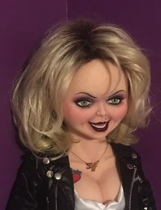 a creepy doll with blonde hair and blue eyes wearing a black leather jacket is posed