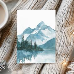 a watercolor painting of a mountain lake surrounded by snow and pine trees with a cup of coffee next to it