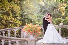 a bride and groom standing on a bridge in front of some trees with orange flowers