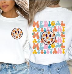 Elevate your medical wardrobe with our Floral Medical Assistant Smiley Sweatshirt. This cozy and stylish sweatshirt features a cheerful floral smiley face design on front, and larger wavy retro graphic on back! This shirt is perfect for any Medical Assistant (MA). Whether you're a Certified Medical Assistant (CMA) or a Registered Medical Assistant (RMA), this graphic tee makes a thoughtful and practical gift for yourself or a fellow assistant.  ♡ DETAILS: .: Medium-heavy fabric .: Loose fit .: S Casual Long Sleeve Nursing Tops, White Long Sleeve Nursing T-shirt, Casual Cotton Sweatshirt For Nursing, White Relaxed Fit Sweatshirt For Nursing, Casual White Nursing Tops, Casual Long Sleeve Nursing T-shirt, Casual Nursing Top With Crew Neck, Casual Nursing Tops With Relaxed Fit, Medical Assistant Outfit