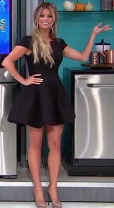 a woman in a short black dress standing next to a refrigerator and smiling at the camera