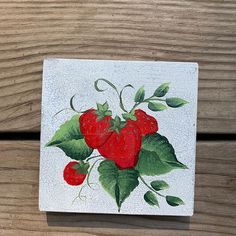 a painting of strawberries with leaves on a white square piece of wood that is sitting on a wooden surface