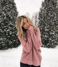 a woman standing in the snow wearing a pink sweater and black pants with her hand on her face