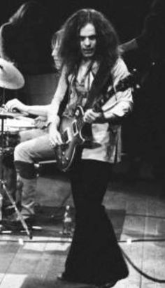 black and white photo of man playing guitar in front of drum kit on stage with other musicians