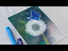 a blue butterfly sitting on top of a dandelion next to two crayons