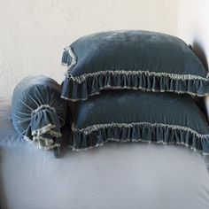 two blue pillows sitting on top of a white bed next to each other with ruffled edges