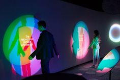 two people standing in front of an art installation with colorful lights on the wall and behind them are silhouettes of people