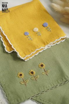 two yellow and green napkins with embroidered flowers on them, next to some feathers