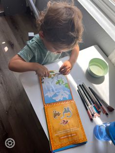 a little boy that is sitting at a table with some paint and pencils on it