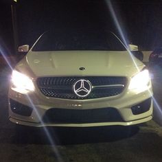 the front end of a white car parked in a parking lot at night with headlights on