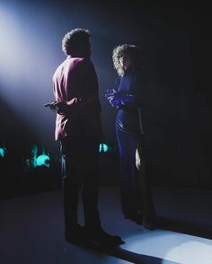 two people standing in the dark with their arms around each other and lights behind them