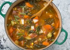 a green pot filled with vegetable soup on top of a marble counter next to a wooden spoon