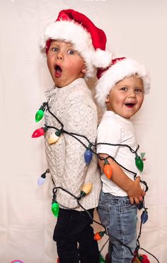 two young boys wearing christmas hats and holding stringed lights on their shoulders, standing in front of a white backdrop