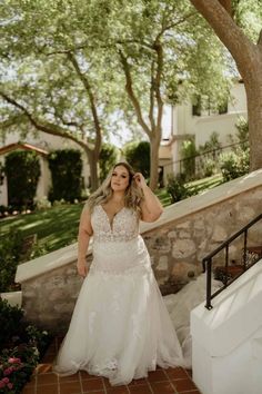 a woman in a wedding dress is standing on stairs near some trees and bushes with her hands behind her head