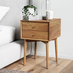 a bedside table with two drawers and a plant on it next to a white bed