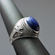 This Sterling Silver Fleur De Lis Cross Lapis Lazuli Oval Gemstone Ring is a stunning blend of elegance and historical charm. Featuring a dark blue lapis lazuli gemstone, known for its protective and intuitive properties, set in a finely crafted sterling silver band. The intricate Fleur De Lis Cross design adds a regal touch, making it perfect for both everyday wear and special occasions. Whether you're treating yourself or looking for a unique gift, this ring is a timeless addition to any jewelry collection. Metal: 925 Sterling Silver (stamped). Lapis Lazuli, with its deep blue hue and golden pyrite flecks, has been prized for centuries for its mesmerizing beauty and spiritual significance. In jewelry, Lapis Lazuli is believed to promote inner peace, clarity of mind, and self-expression. Luxury Lapis Lazuli Oval Rings, Luxury Oval Lapis Lazuli Rings, Classic Round Lapis Lazuli Jewelry, Elegant Blue Oval Cabochon Signet Ring, Elegant Lapis Lazuli Oval Cabochon Jewelry, Elegant Oval Cabochon Lapis Lazuli Jewelry, Blue Lapis Lazuli Gemstone Signet Ring, Blue Lapis Lazuli Signet Ring With Gemstone, Classic Lapis Lazuli Jewelry Ring