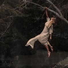 a woman in white dress jumping into water with trees and bushes behind her on the edge of a river