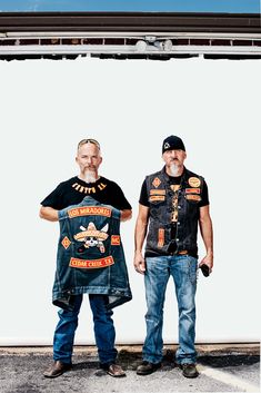 two men standing next to each other in front of a white wall wearing motorcycle vests