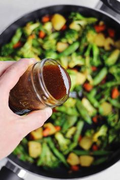 a person holding a jar filled with sauce next to a skillet full of vegetables