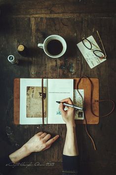 a person writing in a book on top of a wooden table next to a cup of coffee