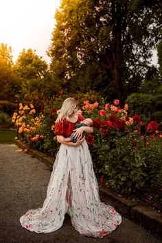 a woman holding a baby in her arms while standing next to some flowers and trees