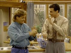 two men standing in a kitchen with one pointing at the other's hand and smiling