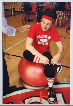 a young man sitting on top of a red ball in a gym with other people around him