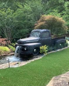 an old black truck is parked in the grass near a stream with water coming out of it