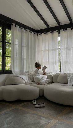 a woman sitting on top of a white couch in front of a window reading a book