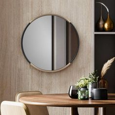 a round mirror hanging on the wall above a wooden table with chairs and vases