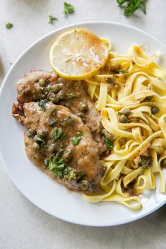 a white plate topped with pasta and meat covered in sauce next to lemon wedges