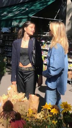 two women are talking to each other in front of some flowers and produce at an outdoor market