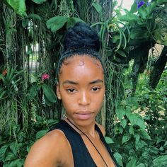 a woman with black hair standing in front of green plants and trees, looking at the camera