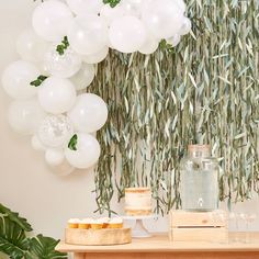 a table topped with white balloons and cake next to a wall covered in greenery