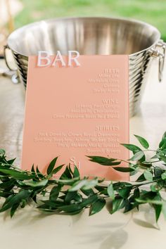 a pink menu sitting on top of a table next to a metal bowl filled with green leaves