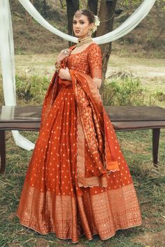 a woman in an orange and white dress standing next to a wooden bench with trees