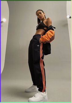 a woman standing in front of a white wall wearing adidas pants and an orange top