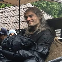 a man with long hair sitting under an umbrella next to another man wearing a black jacket