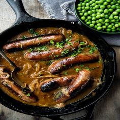 some sausages and noodles in a skillet next to peas on a wooden table