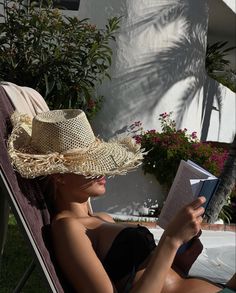 a woman in a straw hat reading a book while sitting on a lawn chair outside