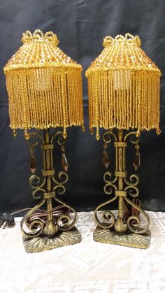 two gold colored lamps with beaded shades on top of each lamp, one is turned off and the other has beads hanging from it