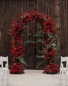 an outdoor ceremony with white chairs and red flowers on the arch in front of a wooden wall