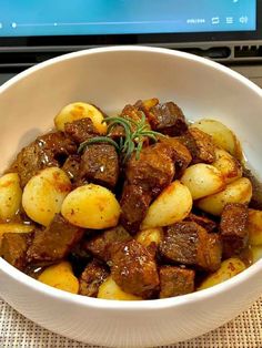 a white bowl filled with meat and potatoes on top of a table next to a laptop